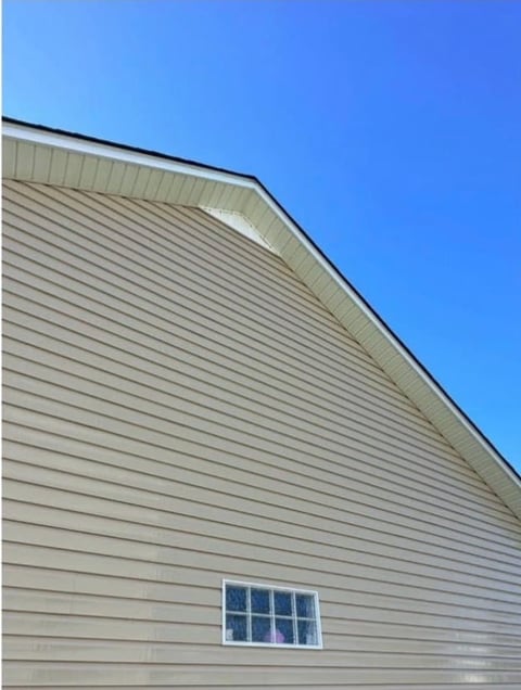 Angled exterior view of a house with light gray vinyl siding and a small window, set against a clear blue sky