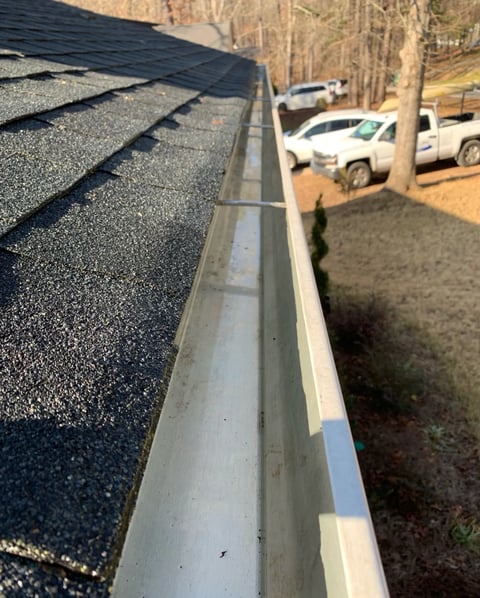 Close-up view of a residential roof edge with asphalt shingles, gutters, and a driveway with parked vehicles below.